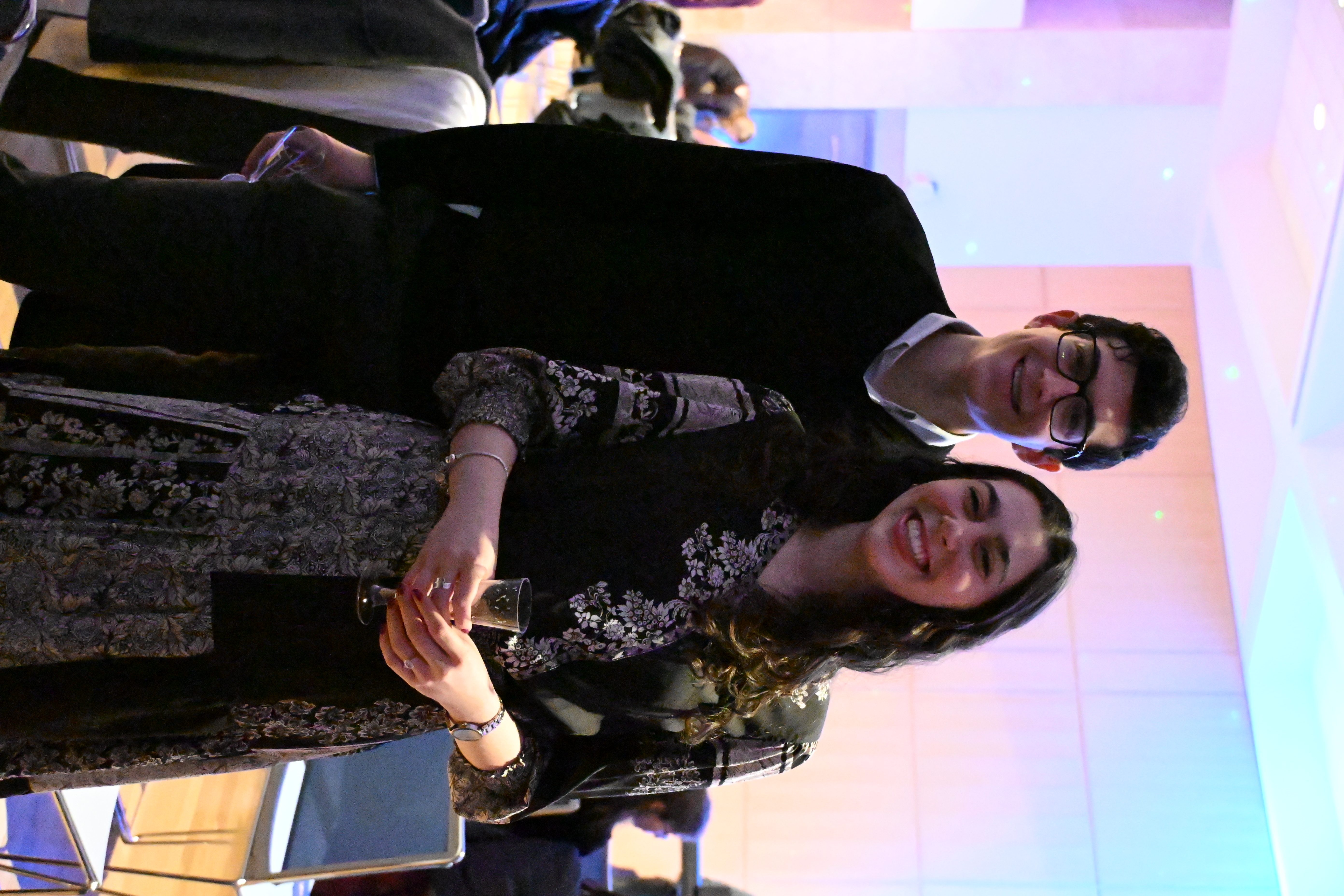 This lively portrait features two young adults, a man and a woman, radiating pure joy with their wide, infectious smiles directed at the camera. The man, in glasses and a black sweater, stands slightly behind the woman, who sports a patterned dark dress and holds a delicate glass, looking utterly delighted. Their coordinated cheerfulness suggests they're having a fantastic time. The background is a soft, colorful blur of pink, blue, and purple lights, creating a festive, almost magical ambiance. This vibrant lighting perfectly sets the scene for a celebratory event, making the whole room feel like it's joining in their merriment. It's a snapshot of genuine happiness, where even the background lights seem to be trying to out-sparkle their smiles. I do not recognize any famous or recognizable content in this image.